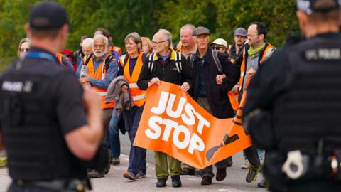 Just Stop Oil protesters approach the gates of Kingsbury oil storage depot, watched by security guards