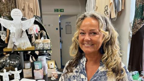 A woman with curly blonde hair looking and the camera and smiling. She is wearing a blue and white top. She is stood in a shop - there are candles to her right