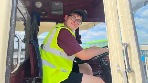 Young man in fluorescent jacket sits in the drivers seat of a bus holding the wheel