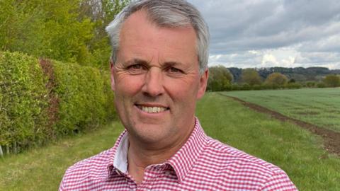 A grey-haired Andrew Ward standing in a field with a hedge to the side. He's wearing a red and white checked shirt and looking towards the camera