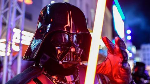 An actor dressed as Darth Vader holds a glowing lightsabre outside a London theatre