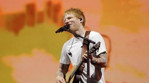 A red-haired Ed Sheeran sings into a microphone while playing a guitar. He is wearing a white tee-shirt
