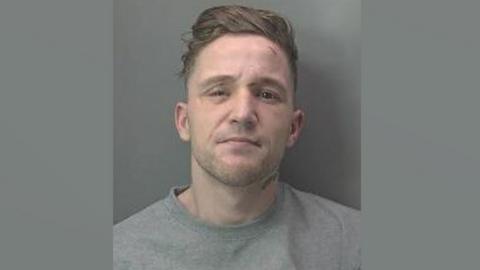 A police mug shot of Dillon Currie aka Abeeb Hussain. He has light brown hair which is longer on the top and very short on the sides and has subble. He is wearing a grey sweatshirt.