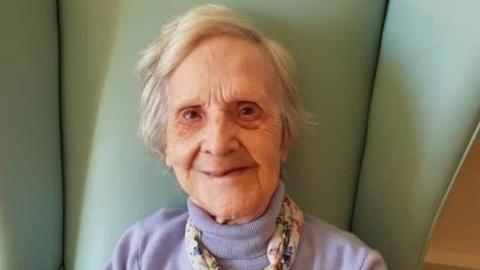 The late Mary ‘Pat’ Thompson smiling and sitting on a green, high-back chair.  She has short, white hair and is wearing a lilac polo-neck jumper and a scarf. 