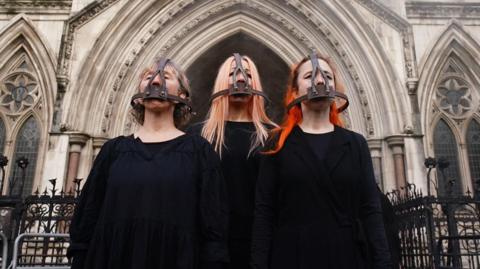 Three campaigners stand in black outside court, wearing metal head-gear which covers their mouths