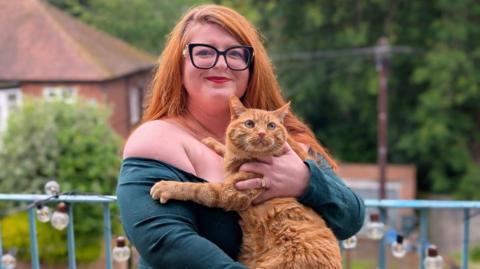 Hayley Hawkins is wearing an off the shoulder green top and holding her ginger cat Simon. A railing behind her has a string of lightbulbs wound through it and there are trees and houses blurred in the background. Hayley is wearing glasses with thick black rims and has very long red hair.
