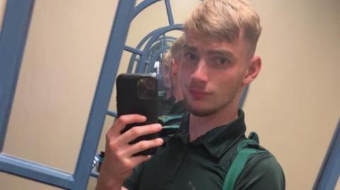 Jamie Kadolski poses for a selfie in a mirror. He is holding up a large phone with a black phone case. He is wearing a green polo neck and slightly smiling at the camera. He has short blonde hair and dark facial hair. 