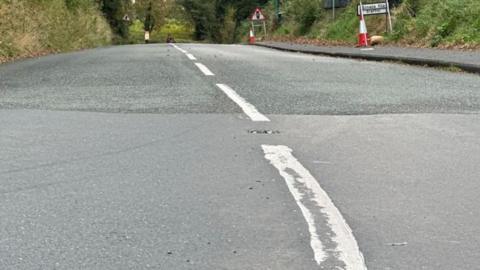 The tarmac and white lines of a roadway, some traffic cones in the distance with a kerb to the right.