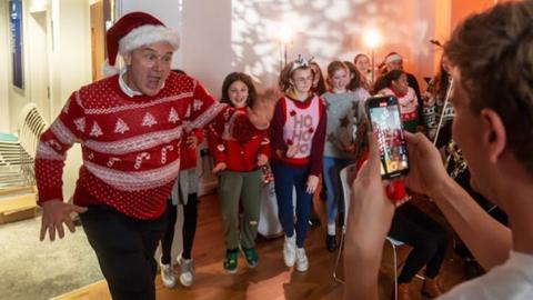 Ed Davey, wearing a Christmas jumper and Santa hat, making a funny face at someone taking a picture on their phone.