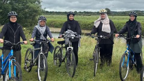 Five women dressed in dark clothing and wearing hijabs and bicycle helmets stand holding five bikes. They are on a grass verge alongside a river, with a grassy field in the background, lined by trees