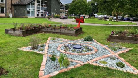 A memorial garden. Flower in wooden planters mark out a square in the grass. In the centre, slate and brick is arranged to a six-pointed Star of David. A plaque is wrapped in red ribbon.