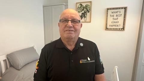 A man stands in a modern-looking bedroom. He has cropped white, thinning hair and wears dark-rimmed glasses and a black polo shirt with the logo "Team Hull 4 Heroes" on it. The bedroom has white walls, a grey wardrobe and a white bed with grey bedding. A picture of a green plant hangs from the wall, while another decoration carries the saying: "What's the best that could happen?"