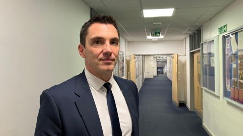 Dominic Wilkes standing in an empty hallway. He is wearing a navy suit jacket with a white shirt and navy tie. The corridor is large behind him and there is a number of signs on the walls.