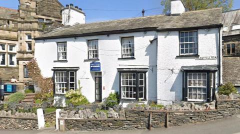 Laurel Cottage is a white cottage with two floors. There is seating and a green area in the front garden. Stone work buildings can be seen on either side.