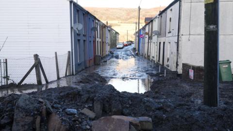 The entrance to a street in daylight is blocked by rubble and mud. Beyond the rubble, water stands in the street and the mountain behind is visible in the background