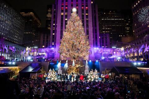 a bright and colourfully lit tree towers over a large group of people standing outside a big building while a small stage in front of the tree is filled with performers