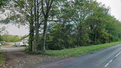 Land behind a row of trees, as seen from a road. There is a single storey building, a white van and a white car seen through a gap in the trees on the land.