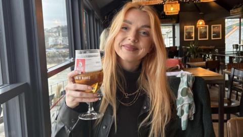 Close up image of  Briony Watkins, 24. The image shows Briony smiling while holding a pint glass in a pub or restaurant.