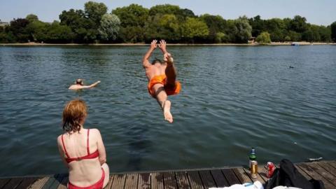 A man backflips into a river from a pontoon, wearing swim shorts. A woman sits on the pontoon with her back to the camera, wearing a bikini