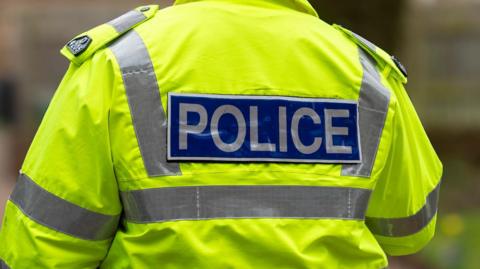 A close-up shot of the back of a police officer, showing his jacket. It is fluorescent yellow, with reflective grey stripes and a large blue panel which reads POLICE in grey lettering.