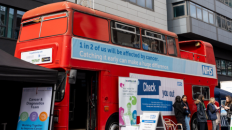 A red double-decker bus with words on the outside including 1 in 2 of us will be affected by cancer. Some people, a chair and signs are outside the bus.  