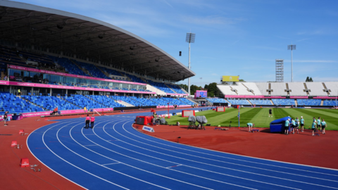 A view of an athletics stadium with a blue running track, green field and spectators under a clear sky.