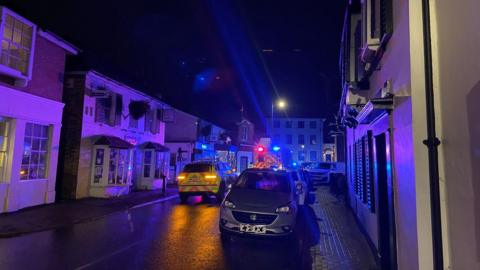 A night time scene showing a village road with two ambulance vehicles parked in the middle of it and a parked car on the side of the road. The ambulance has its red and blue lights lit up but the street is otherwise dark