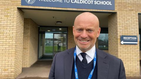 Alun Ebenezer wears a suit and a lanyard, smiling at the camera in front of a sign saying Welcome to Caldicot school
