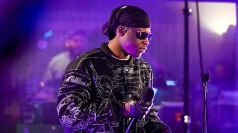 A man with a black head covering, sunglasses and a long-sleeved black jacket with gloves, holds a microphone as he looks to the side while performing on stage.