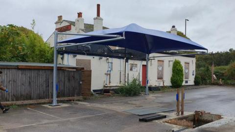 The former cafe rouge building seen from behind and with a blue awning installed on a metal stand over a car park. The building is boarded up and a hole has been due in the ground in the car park.