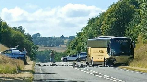 A yellow and brown bus on the side of the road. The left hand side of the bus is damaged, with debris on the road. A white van can be seen on the other side of the road. A person stands in the middle of the road, with two vehicles in the distance. 
