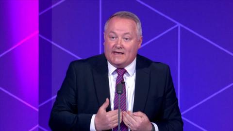 Darren Miller from Welsh Conservatives is standing in front of a purple background. Darren Miller is wearing a black blazer with a white shirt and a blue and red stripy tie.
