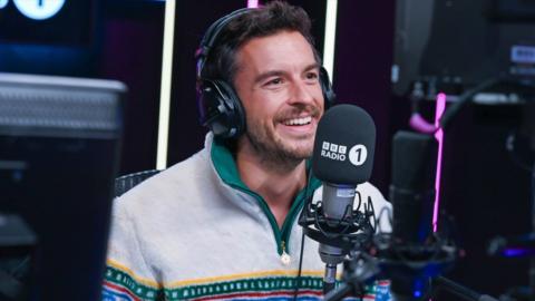 Jonathan Bailey smiling in front of a Radio 1 branded microphone and headphones in the studio.