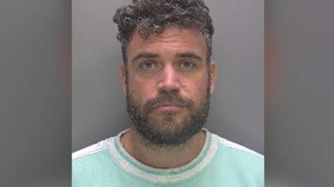 Custody photo of Nathan King stood against a grey background. He is wearing a light blue top with a white line around the neck of the shirt. He has dark curly hair and a beard. 