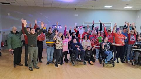 Around 20 people supported by the Ark Trust pose for a photo during a Christmas disco. Some are wheelchair users and most are wearing Christmas jumpers or hats. Disco lights can be seen on the walls