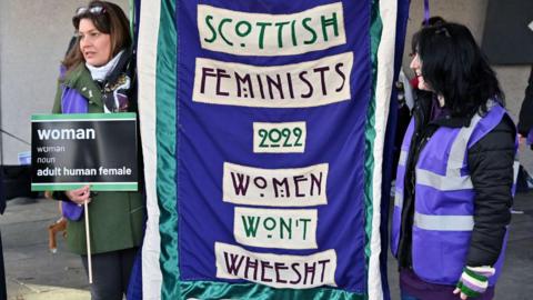 Two women holding up a purple banner with the words 'Scottish Femenists Women Won't Wheesht' on the front in white boxes.