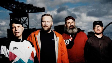 The rock band Mogwai pose against a stormy sky in an industrial landscape