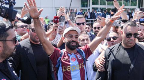 Mohamed Salah, visible from stomach up wearing a white baseball cap and Trabzonspor home shirt featuring wide vertical sky blue and claret stripes, smiles as he raises both arms up in the air as a greeting to watching fans who are not in shot but have gathered at Istanbul Airport. Salah is surrounded by several large male bouncers in black suits and dark glasses while members of the media also jostle, waving phones, microphones and cameras