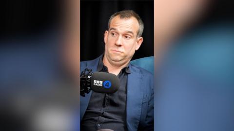 A man with short grey hair wearing a black shirt and blue blazer raises his eyebrows at someone off-camera