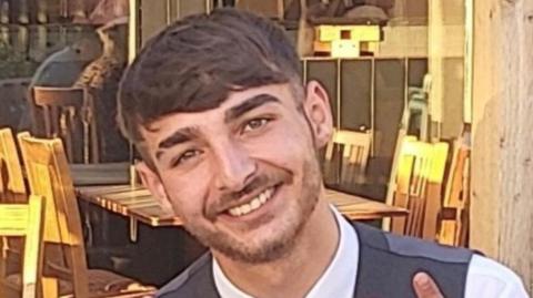 Mohammed Shaan Ali Rasul, with brown hair, brown eyes and brown stubble is smiling at the camera. There are tables and chairs in the background of what appears to be an outdoor seating area.