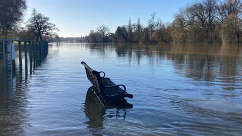 An empty half-submerged bench sits surrounded by water in a flooded river scene. A line of trees can be seen in the background amidst clear blue sunny skies.