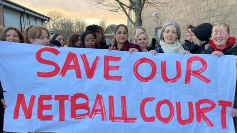 A group of women holding up a big white sign that says 'Save our netball court' in red writing. They are stood outside a leisure centre. 