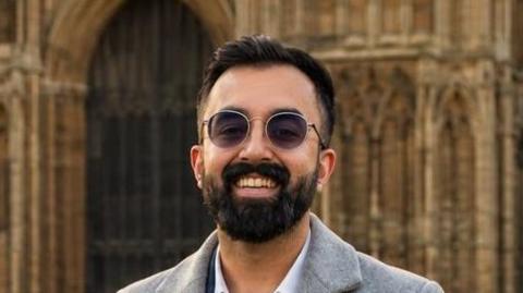 A man smiling straight at the camera while standing outside a cathedral. He is wearing a grey coat over a white shirt and has sunglasses on.
