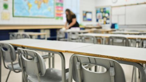 Grey chairs at desks sitting in rows in the classroom of a school with a female teacher working in the background. She is wearing a black topped and is blurred. There is a map of the world on the wall.