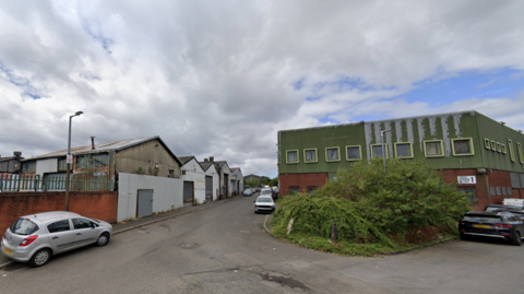 Industrial units sit on one side of the road with a large green and brick building on the right hand side. Cars are parked on both sides of the street which widens out at the bottom of the image.