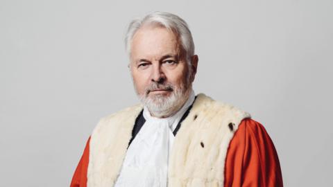 Sir Timothy Le Cocq wearing traditional court dress. He is has grey hair and a grey short beard. He is wearing a red cloak with a white fur finish 