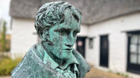 Weathered looking greenish statue of John Clare. Its head is turning sideways and looking down, has deep eyes and sideburns. Behind it is a low-lying white painted building with black painted windows.