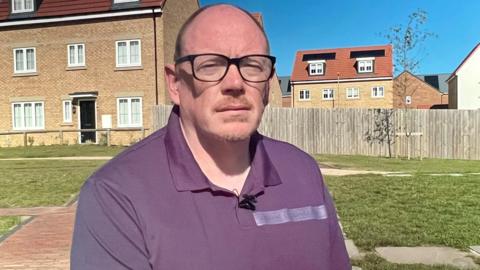 A man in his fifties is sitting on a park bench in front of a row of new-build houses.  He is wearing a purple polo shirt and black framed glasses.  It is a sunny day with blue sky.