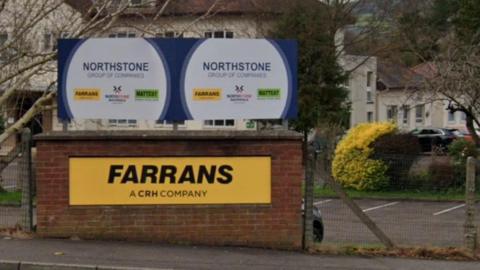 The company name Farrans is displayed in black letters on a yellow background which is attached to a red brick wall. There are buildings and a parking lot in the background
