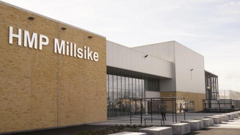 The front of a modern prison with a high brick wall giving way to a modern-looking frontage made up of black glass and grey cladding. A sign in large white letters reads "HMP Millsike".